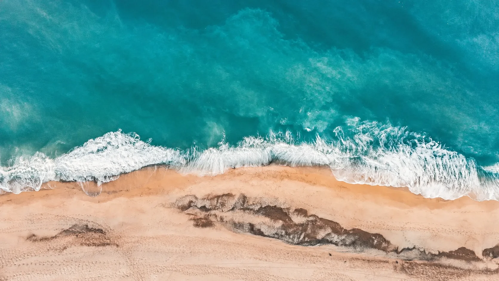 Blue-green waves meeting a sunlit shoreline, viewed from above